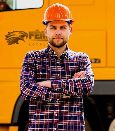 imagem com um profissional da construção, de epi, com braços cruzados, em frente ao caminhão da fênix, com cabine laranja
